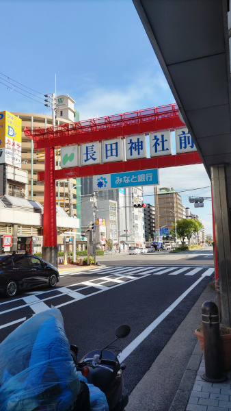 道路に建っている「長田神社前」の大きな鳥居。朱色の鳥居に白の看板、青文字で「長田神社前」と書いてあり、その下にはなんのみなと銀行の看板が下がっています