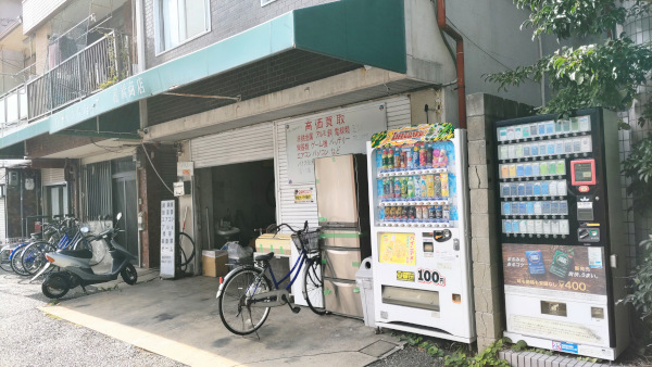 リサイクルショップの写真です。緑色の屋根で店先に売り物らしく冷蔵庫が置かれています。